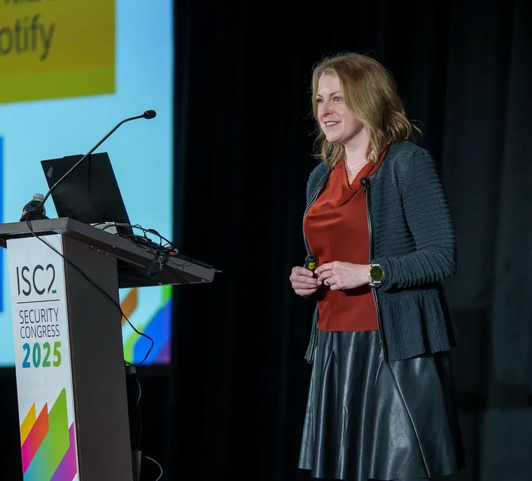 Lora Vaughn smiling, holding a presentation clicker, speaking on stage at a branded ISC2 Security Congress 2025 lectern